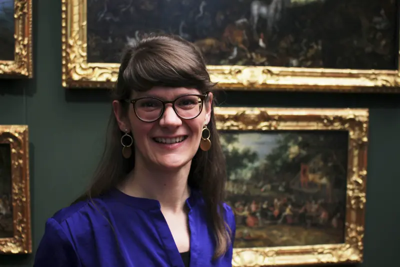 Eine Frau schaut im Kameraobjektiv. Hinter ihr sind verschiedene Gemälde zu sehen. 